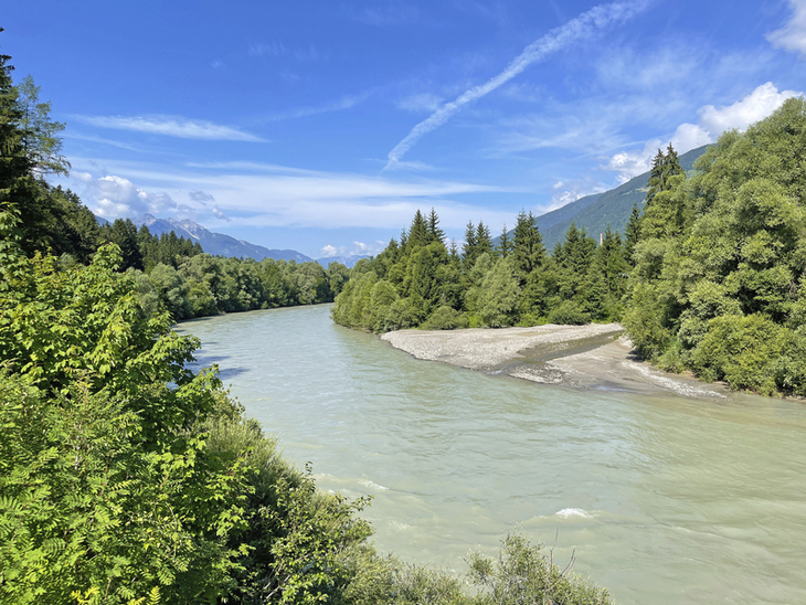Fluss von Bäumen gesäumt, Berge im Hintergrund, blauer Himmel mit Wolken.