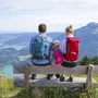 Familie auf Bank mit Wanderrucksäcken, blickt auf Berge und See im Salzkammergut.