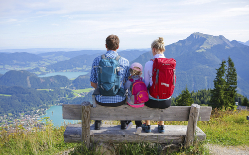 Familie auf Bank mit Wanderrucksäcken, blickt auf Berge und See im Salzkammergut.