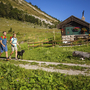 Zwei Personen und ein Hund wandern an einer Almhütte im Salzkammergut vorbei.