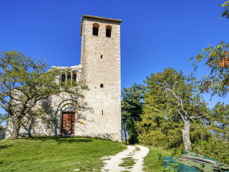 Kirche San Giuliano bei Spoleto, umgeben von Bäumen, klarer blauer Himmel.