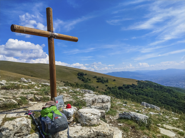 Holzkreuz auf einem Hügel mit Wanderausrüstung im Vordergrund und weiter Aussicht.