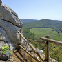 Wanderpfad mit Rucksack und Wanderstöcken, Blick auf grüne Hügel und Felsen.