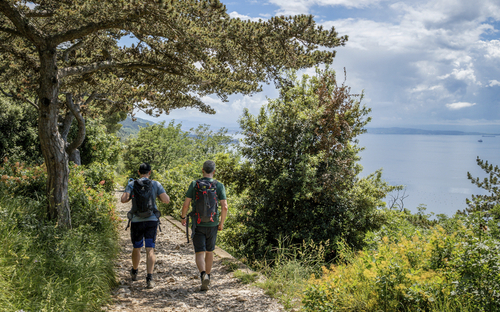 Zwei Wanderer auf einem Pfad in einer grünen Landschaft mit Blick auf das Meer.
