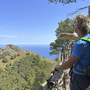 Person wandert auf einem Pfad mit Blick auf bewaldete Hügel und das Meer.
