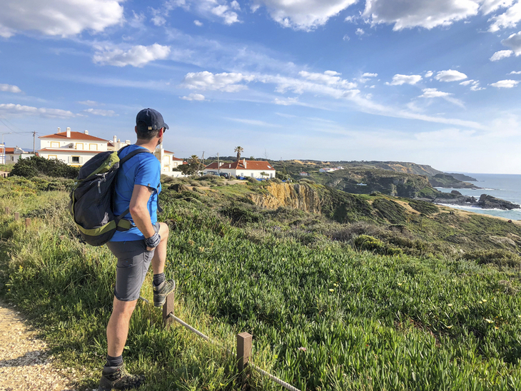 Wanderer blickt auf Küstenlandschaft mit Häusern und Meer im Hintergrund.