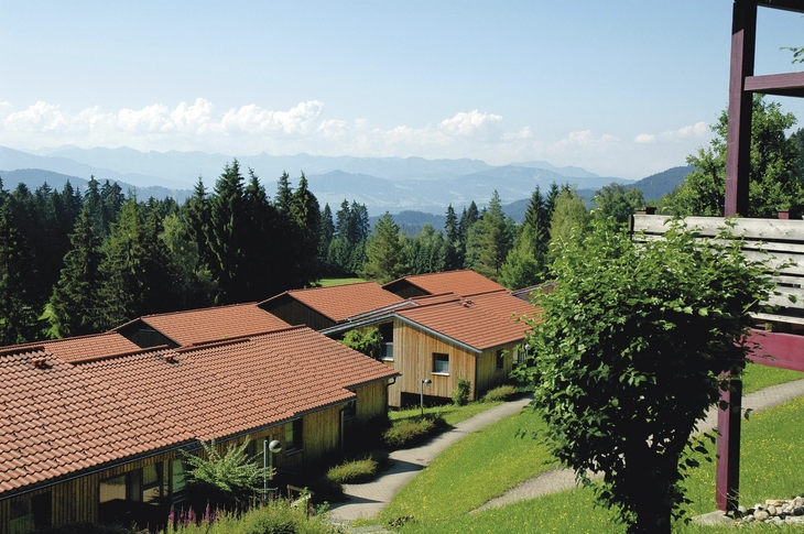 Häuser mit roten Dächern vor Alpenpanorama und bewaldeter Landschaft.