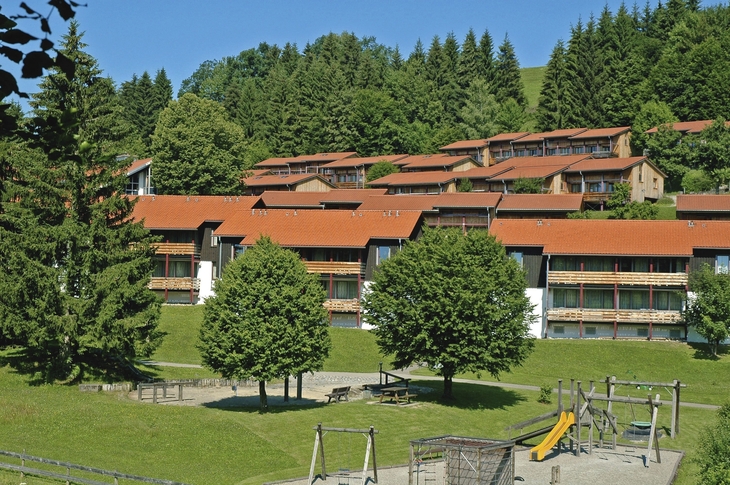 Außenansicht eines Gebäudekomplexes im Grünen mit Spielplatz im Vordergrund.