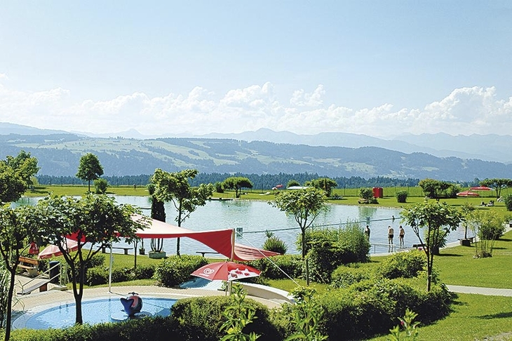Alpenfreibad in Landschaft mit Bergen, Bäumen und Sonnenschirmen.