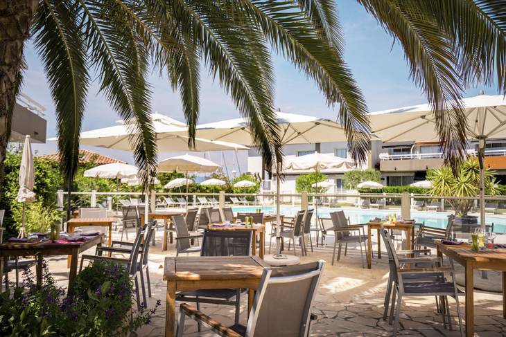 Terrasse eines Restaurants mit Tischen und Stühlen, Sonnenschirmen, Palmen, Pool im Hintergrund.