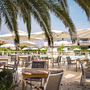 Terrasse eines Restaurants mit Tischen und Stühlen, Sonnenschirmen, Palmen, Pool im Hintergrund.