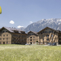 Drei Gebäude des Dormio Aparthotel Hinterstoder vor Bergkulisse im Sommer; Gleitschirmflieger.