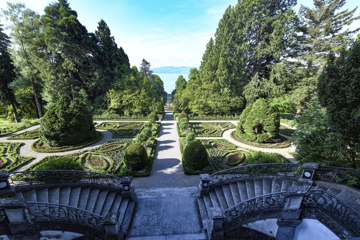 Blick von der Treppe der Villa Alwind, in der Ferienanlage Lindau, auf einen symmetrisch angelegten historischen Garten mit Hecken und Wegen vor Bäumen. Dahinter sieht man den Bodensee unter leichtbewölktem blauen Himmel.