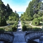 Blick von der Treppe der Villa Alwind, in der Ferienanlage Lindau, auf einen symmetrisch angelegten historischen Garten mit Hecken und Wegen vor Bäumen. Dahinter sieht man den Bodensee unter leichtbewölktem blauen Himmel.
