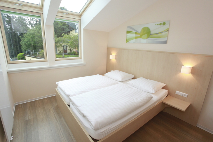 Helles Schlafzimmer in der Ferienanlage Lindau, im Haus an der Obstwiese, mit Doppelbett mit Lampen, senkrechten Fenstern und Dachfenstern und einem Bild über dem Bett. .