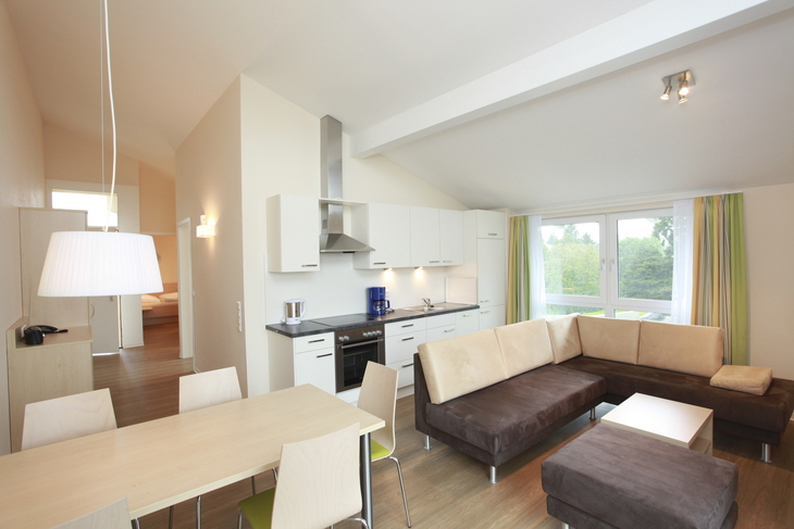 Wohnzimmer im Appartementhaus an der Obstwiese, in der Ferienanlage Lindau, mit heller Küchenzeile, Esstisch mit Stühlen, Ecksofa, kleinem Tisch und großem Fenster mit Vorhängen.