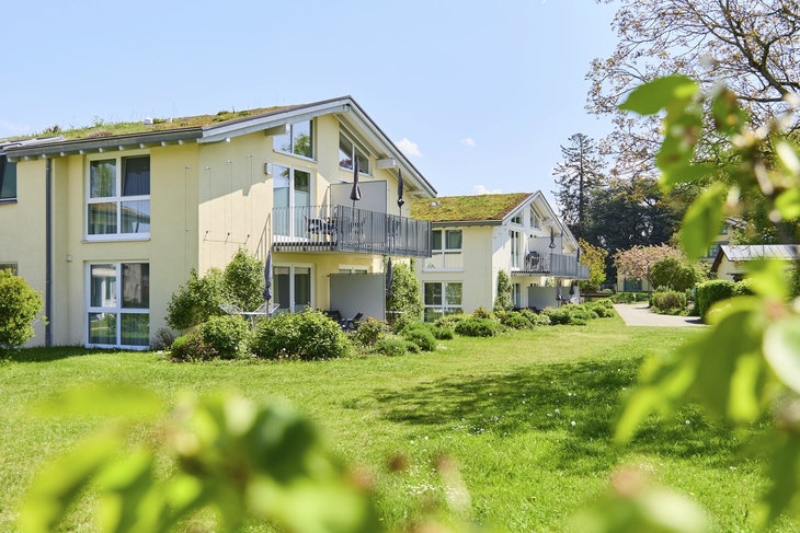 Hellgelbe Appartementhäuser an der Obstwiese, der Ferienanlage Lindau, mit Balkonen auf einer grünen Rasenfläche und Bäumen unter blauem Himmel.