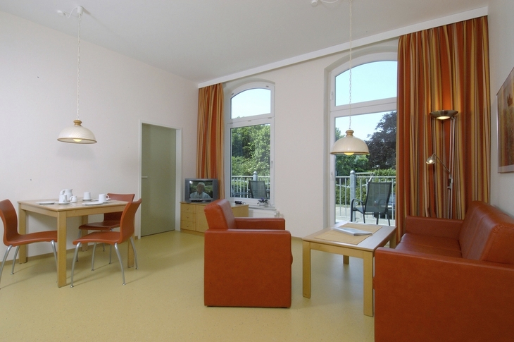 Wohnzimmer mit Esstisch, roten Sesseln, Sofa, Fernseher und großen Fenstern mit Blick nach außen.