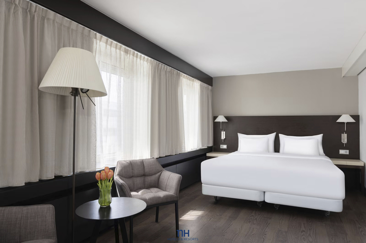 Hotelzimmer mit Doppelbett, Sesseln, Tisch, Stehlampe und Fenster mit Vorhängen.