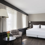 Hotelzimmer mit Doppelbett, Sesseln, Tisch, Stehlampe und Fenster mit Vorhängen.