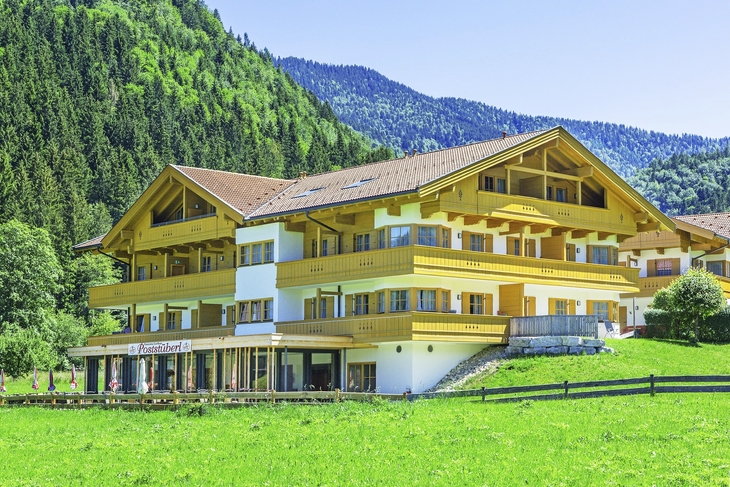 Gelbes Gebäude vor bewaldetem Hügel mit Wiese im Vordergrund.