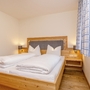Schlafzimmer in der Ferienanlage Oberwössen, mit Holzfußboden, einem Doppelbett mit Nachttischen und Nachttischlampen sowie einem Fenster mit blau-weiß karierten Vorhängen.