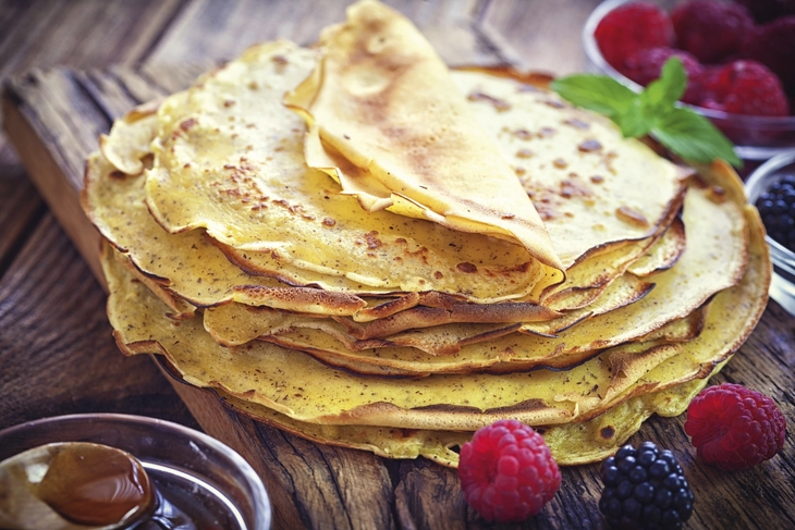 Ein Stapel Crêpes auf einem Holzbrett mit Beeren im Hintergrund.