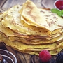 Ein Stapel Crêpes auf einem Holzbrett mit Beeren im Hintergrund.
