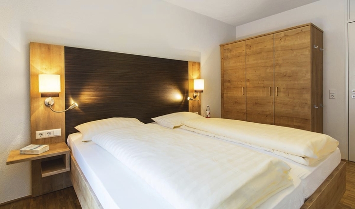 Gemütliches Schlafzimmer in der Ferienanlage Titisee mit Doppelbett mit hohem Kopfteil, Nachttischen und Lampen und einem dreitürigen Holzschrank.