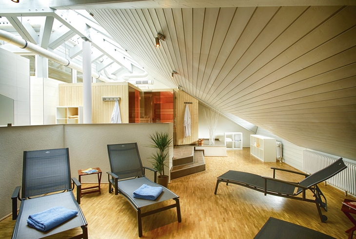 Moderner Entspannungsraum mit drei Liegestühlen auf Holzboden, unter der Dachschräge mit Holzverkleidung und Glasdach, in der Ferienanlage Titisee. Im Hintergrund die beiden Saunaräume und Bademäntel an den Hakenleisten daran.