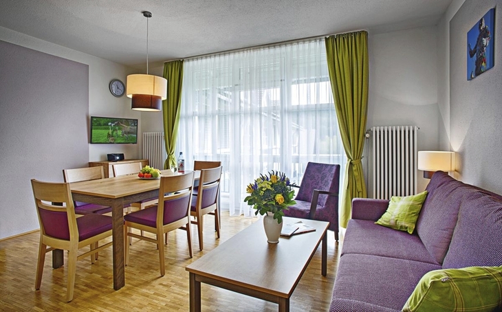 Wohnbereich der Ferienanlage Titisee mit Holzfußboden, Esstisch und Stühlen aus Holz, sowie einem lila Sofa und Sessel mit kleinem Holztisch davor, auf dem eine Vase mit Blumen steht. Links, vor dem großen Fenster mit weißer Gardine und grünem Vorhang, hängt ein Flachbildschirm an der Wand über einem Sideboard unter einer Wanduhr.