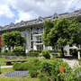 Weißes, mehrstöckiges Gebäude der Ferienanlage Titisee unter leicht bewölktem blauen Himmel. Im Vordergrund der Garten mit einer Parkbank, Wiese, Bäumen und einem gepflasterten Weg zum Gebäude.