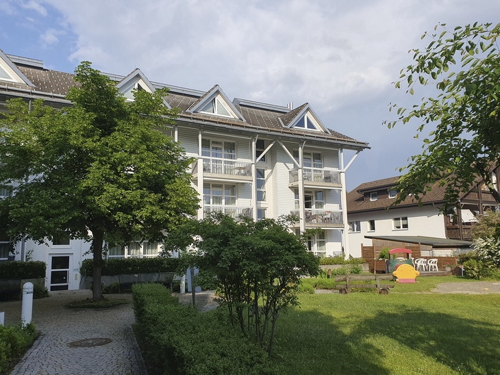 Weißes, mehrstöckiges Gebäude der Ferienanlage Titisee unter leicht bewölktem blauen Himmel. Im Vordergrund der Garten mit Wiese, Bäumen und einem gepflasterten Weg zum Gebäude.