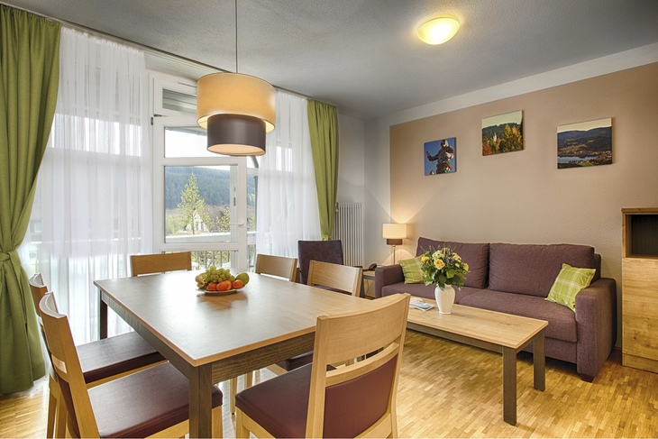 Wohn-Essbereich in der Ferienanlage Titisee, mit Holzfußboden, Esstisch mit sechs Stühlen, Sofa und Sessel mit kleinem Tisch, großer Fensterfront mit Gardine und Vorhängen sowie einer Tür auf den Balkon.