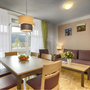 Wohn-Essbereich in der Ferienanlage Titisee, mit Holzfußboden, Esstisch mit sechs Stühlen, Sofa und Sessel mit kleinem Tisch, großer Fensterfront mit Gardine und Vorhängen sowie einer Tür auf den Balkon.