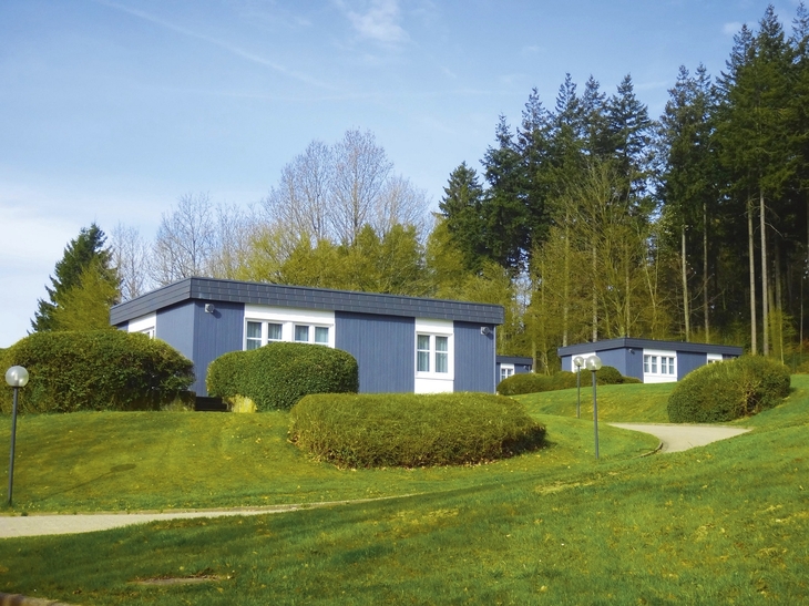 Mehrere blaue Bungalows mit Flachdach und weißen Fenstern, der Ferienanlage Prüm auf einer grünen Wiese mit Wald im Hintergrund.