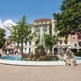 Brunnen auf der Piazza Manzoni in Lugano, umgeben von bunten Gebäuden.