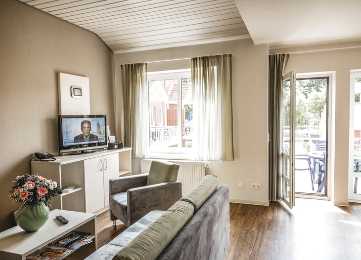 Wohnbereich in der Ferienanlage Büsum, mit Sofa, Sessel, kleinem Tisch und Fernseher auf einem Sideboard, sowie einem Fenster und Tür zum Balkon.