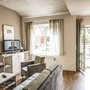 Wohnbereich in der Ferienanlage Büsum, mit Sofa, Sessel, kleinem Tisch und Fernseher auf einem Sideboard, sowie einem Fenster und Tür zum Balkon.