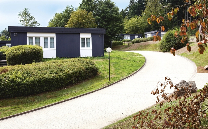 Anthrazitfarbene Bungalows mit Flachdach und weißen Fenstern und Türen, der Ferienanlage Prüm, an einem gewundenen Weg in einer grünen, bewaldeten Umgebung.