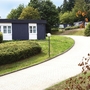 Anthrazitfarbene Bungalows mit Flachdach und weißen Fenstern und Türen, der Ferienanlage Prüm, an einem gewundenen Weg in einer grünen, bewaldeten Umgebung.