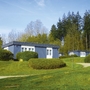 Mehrere blaue Bungalows mit Flachdach und weißen Fenstern, der Ferienanlage Prüm auf einer grünen Wiese mit Wald im Hintergrund.