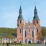 Basilika in Prüm mit zwei Türmen und barocker Fassade unter blauem Himmel vor Bäumen.