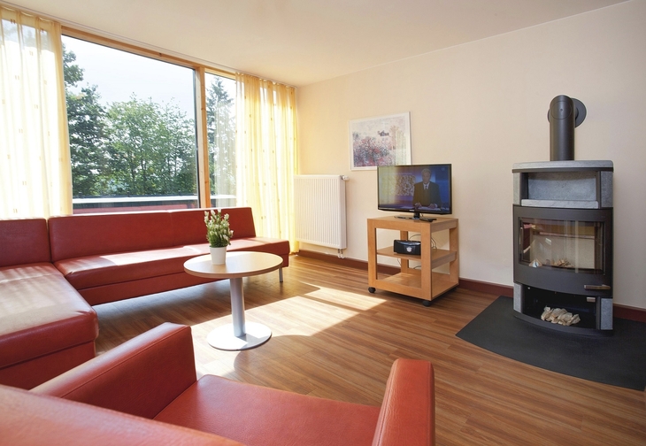 Wohnzimmer in der Ferienanlage Prüm, mit rotem Ecksofa und Sessel, kleiner runder Tisch, Holzofen, Fernseher und Blick auf Bäume durch großes Fenster mit gelben Vorhängen rechts und links.