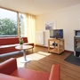Wohnzimmer in der Ferienanlage Prüm, mit rotem Ecksofa und Sessel, kleiner runder Tisch, Holzofen, Fernseher und Blick auf Bäume durch großes Fenster mit gelben Vorhängen rechts und links.