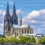 Kölner Dom vor blauem Himmel mit Wolken und grünen Bäumen im Vordergrund.