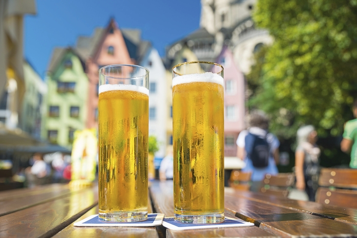 Zwei Gläser Kölschbier auf einem Holztisch vor bunten Häusern in Köln.