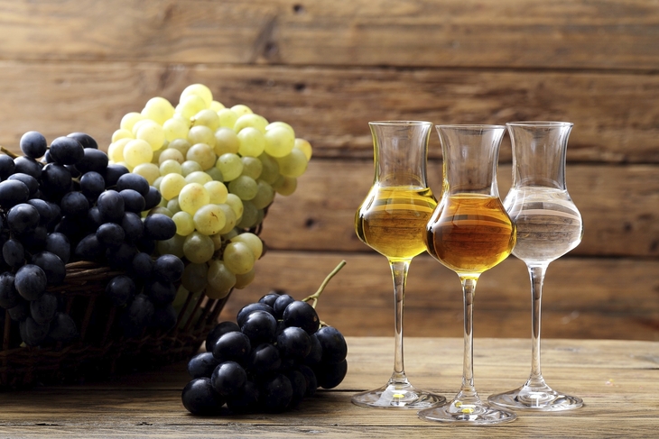 Drei Gläser Grappa mit Trauben im Hintergrund auf Holzuntergrund.