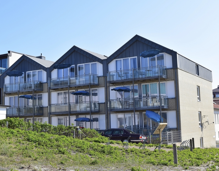 Dreistöckige aneinander gebaute Häuser mit spitzen Dächern, der Ferienanlage Norderney, mit Balkonen, ausgestattet mit Sitzmöbeln und blauen Sonnenschirmen vor klarem Himmel und grünem Bewuchs im Vordergrund.