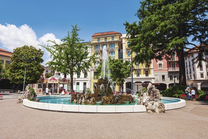 Brunnen auf der Piazza Manzoni in Lugano, umgeben von bunten Gebäuden.